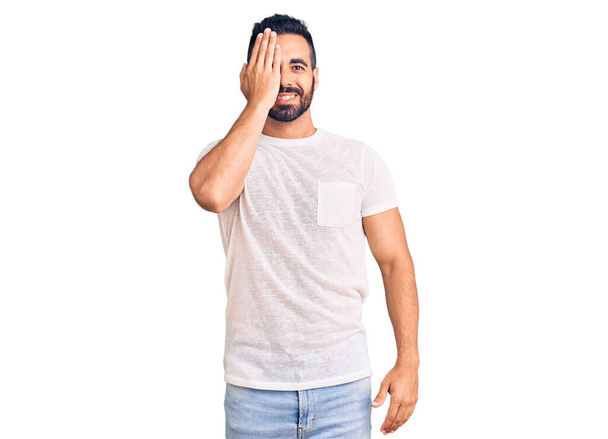 Young hispanic man wearing casual clothes covering one eye with hand, confident smile on face and surprise emotion.  - 写真・画像