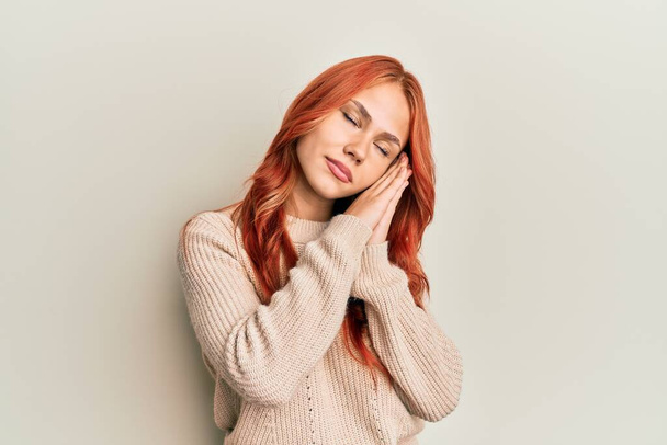 Junge schöne rothaarige Frau trägt lässigen Winterpullover schlafen müde träumend und posiert mit den Händen zusammen, während sie mit geschlossenen Augen lächelt.  - Foto, Bild