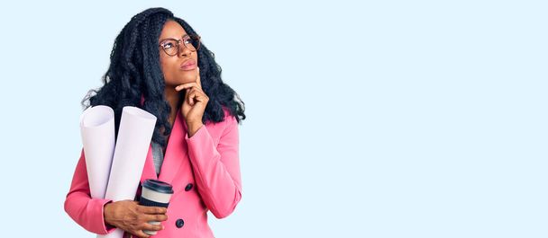 Όμορφη Αφροαμερικανή γυναίκα που κρατάει χάρτινα σχέδια και πίνει καφέ σοβαρό πρόσωπο σκεπτόμενη την ερώτηση με το χέρι στο πηγούνι, σκεπτόμενη την μπερδεμένη ιδέα  - Φωτογραφία, εικόνα