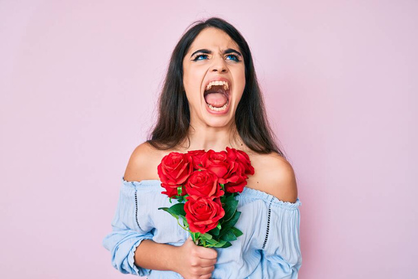 Brunette tiener meisje met bloemen boos en gek geschreeuw gefrustreerd en woedend, schreeuwen van woede opkijken.  - Foto, afbeelding