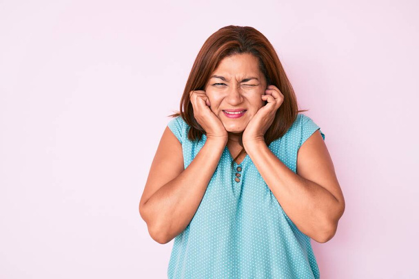 Brünette hispanische Frau mittleren Alters in lässiger Kleidung bedeckt Ohren mit Fingern mit genervtem Gesichtsausdruck wegen des Geräusches lauter Musik. Gehörloses Konzept.  - Foto, Bild