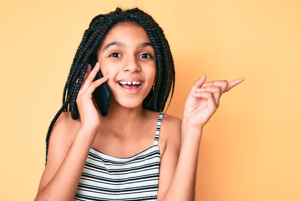 Junge afrikanisch-amerikanische Mädchen mit Zöpfen unterhalten sich auf dem Smartphone, lächeln glücklich und zeigen mit Hand und Finger zur Seite  - Foto, Bild