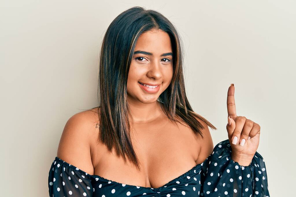 Joven chica latina usando ropa casual sonriendo con una idea o pregunta apuntando con el dedo hacia arriba con la cara feliz, número uno  - Foto, imagen