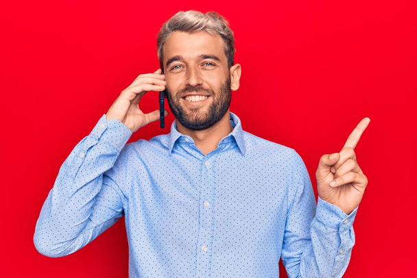 Jonge knappe blonde man met baard hebben gesprek praten op de smartphone lachend gelukkig wijzend met hand en vinger naar de zijkant - Foto, afbeelding