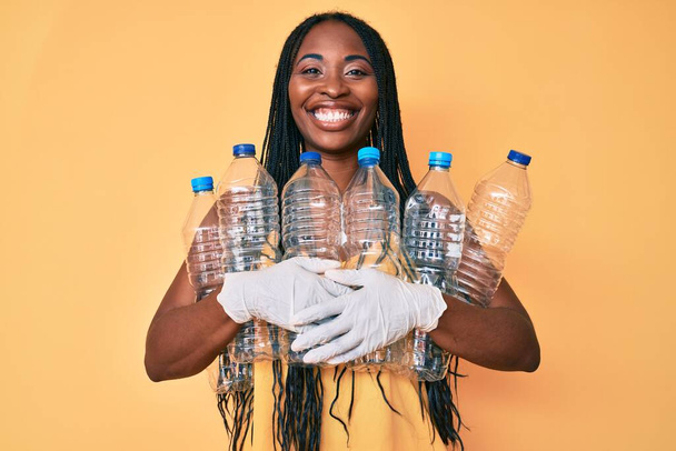 Femme afro-américaine avec des tresses tenant des bouteilles en plastique recyclables souriant avec un sourire heureux et cool sur le visage. montrant les dents.  - Photo, image