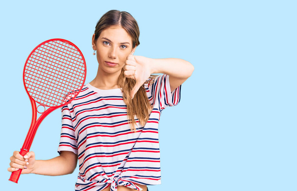 Όμορφη καυκάσια γυναίκα με ξανθά μαλλιά παίζει τένις κρατώντας ρακέτα με θυμωμένος πρόσωπο, αρνητική ένδειξη δείχνει αντιπάθεια με τους αντίχειρες προς τα κάτω, την έννοια της απόρριψης  - Φωτογραφία, εικόνα