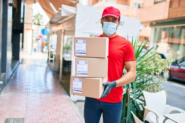 Üniforma giyen ve Coronavirus koruması takan genç bir teslimatçı. Sağlık maskesi. Kasaba sokaklarında karton paketler tutuyor.. - Fotoğraf, Görsel