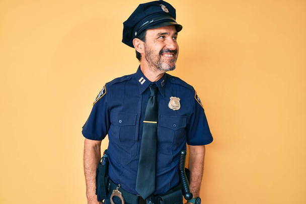 Hispanischer Mann mittleren Alters in Polizeiuniform, der mit einem Lächeln im Gesicht und natürlichem Gesichtsausdruck zur Seite schaut. Lachen selbstbewusst.  - Foto, Bild