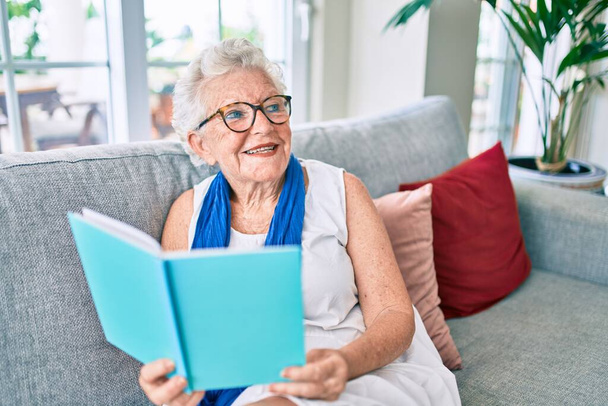 Ηλικιωμένη ηλικιωμένη γυναίκα με γκρίζα μαλλιά χαμογελώντας χαρούμενη καθισμένη στον καναπέ διαβάζοντας ένα βιβλίο στο σπίτι - Φωτογραφία, εικόνα