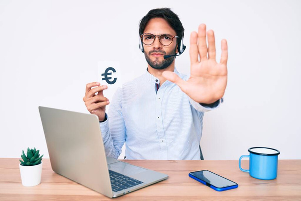Schöner hispanischer Mann im Büro mit Euro-Banner in der Hand und Stoppschild mit ernstem und selbstbewusstem Gesichtsausdruck, Verteidigungsgeste  - Foto, Bild