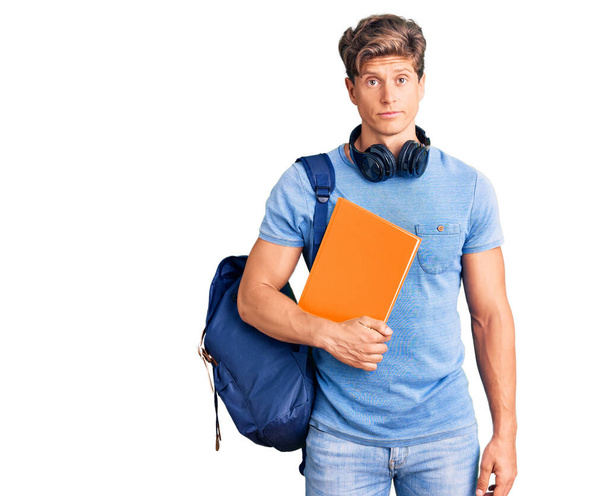 Mladý pohledný muž nosí studentský batoh a sluchátka drží knihu myšlení postoj a střízlivý výraz hledá sebevědomí  - Fotografie, Obrázek