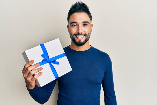 Young man with beard holding gift looking positive and happy standing and smiling with a confident smile showing teeth  - 写真・画像