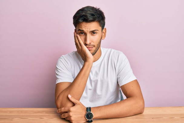 Young handsome man wearing casual white t-shirt sitting on the table thinking looking tired and bored with depression problems with crossed arms.  - Photo, Image