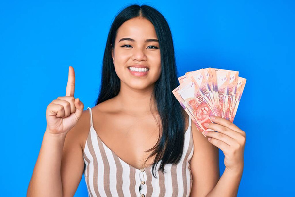 Young beautiful asian girl holding south african rand banknotes smiling with an idea or question pointing finger with happy face, number one  - Foto, Imagen