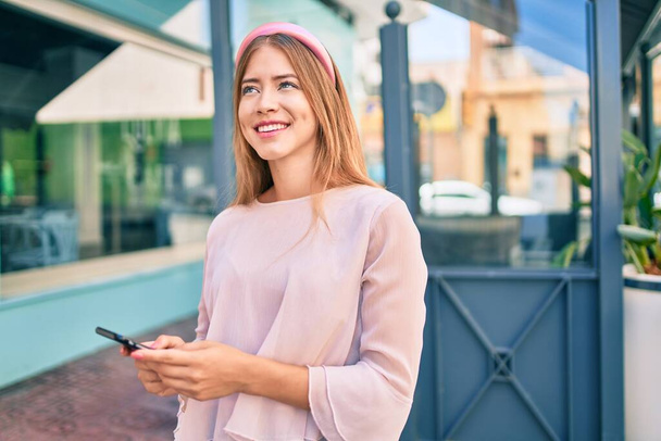 Young caucasian girl smiling happy using smartphone at the city. - 写真・画像