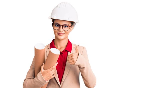 Young beautiful woman wearing architect hardhat and glasses holding blueprints smiling happy and positive, thumb up doing excellent and approval sign  - Valokuva, kuva