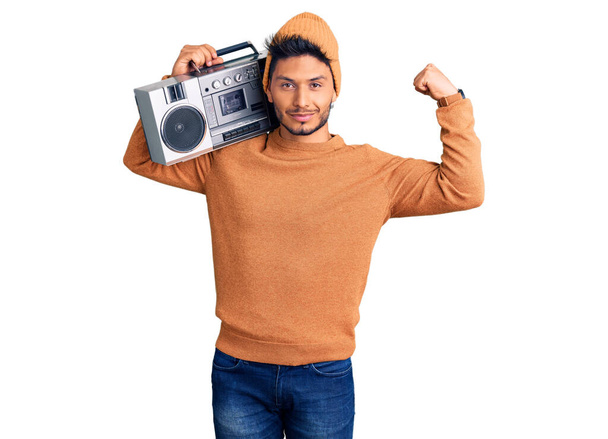 Guapo joven latinoamericano sosteniendo boombox, escuchando música fuerte persona mostrando el músculo del brazo, confiado y orgulloso de poder  - Foto, imagen
