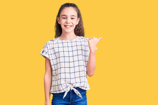 Cute latynoski dziewczynka noszenie ubrań casual uśmiecha się z szczęśliwy wygląd twarzy i wskazując na stronie z kciukiem w górę.  - Zdjęcie, obraz