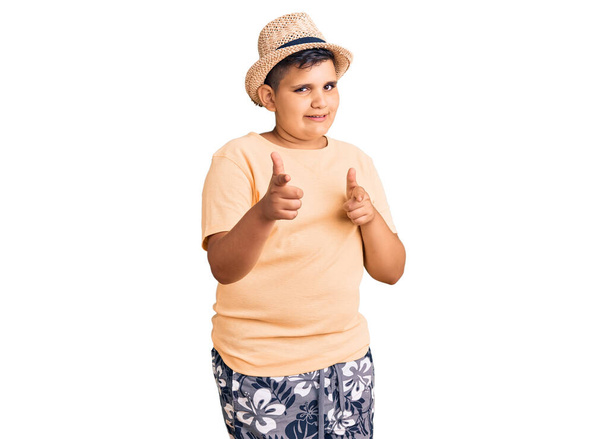 夏の帽子とハワイの水着を身に着けている小さな男の子は、幸せで面白い顔をしたカメラに指を指しています。良いエネルギーと振動は.  - 写真・画像