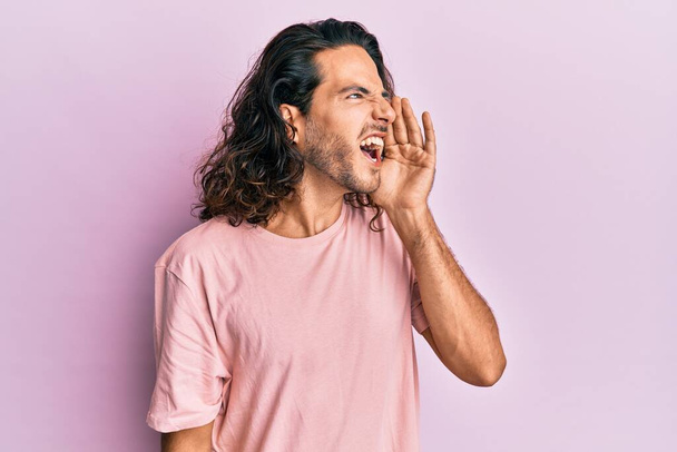 Mladý pohledný muž s dlouhými vlasy, oblečený do neformálního oblečení, křičící a křičící na stranu s rukou na ústech. komunikační koncept.  - Fotografie, Obrázek