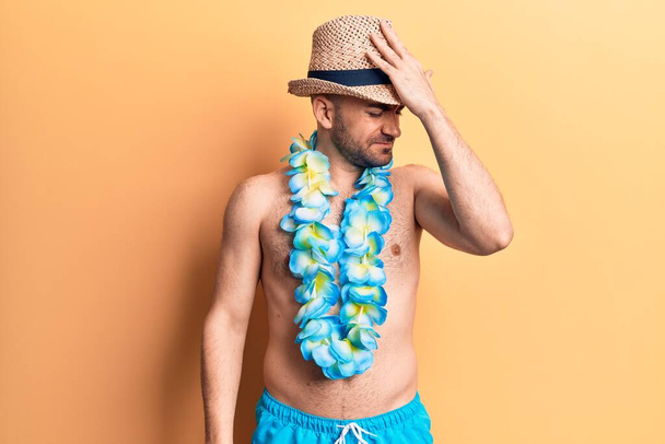 Junger gutaussehender, glatzköpfiger Mann in Badebekleidung und hawaiianischem Lei überrascht mit der Hand auf dem Kopf für Fehler, erinnere dich an Fehler. Vergessenes, schlechtes Gedächtnis.  - Foto, Bild