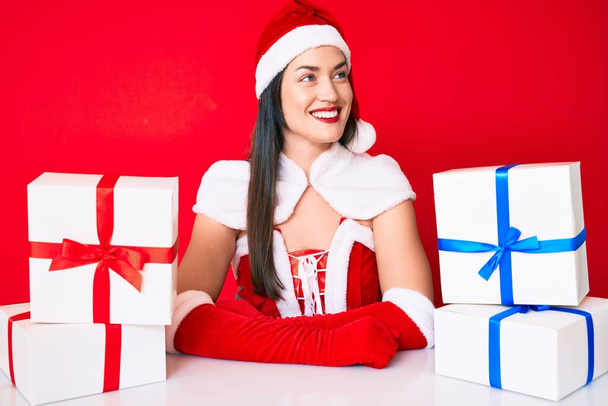 Mujer joven con traje de Santa Claus sentado en el escritorio con regalos mirando a un lado, pose de perfil relajante con rostro natural y sonrisa confiada.  - Foto, Imagen