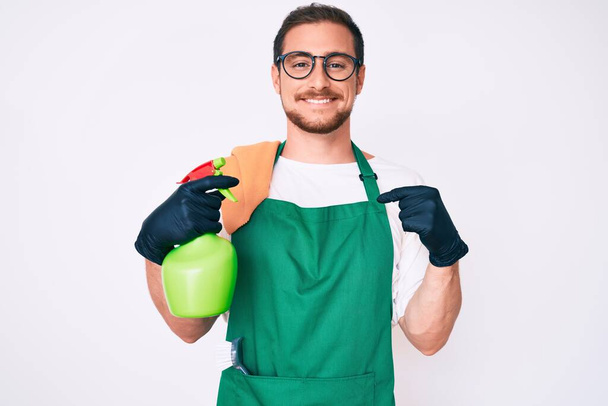 Young handsome man wearing apron holding sprayer pointing finger to one self smiling happy and proud  - Foto, Bild