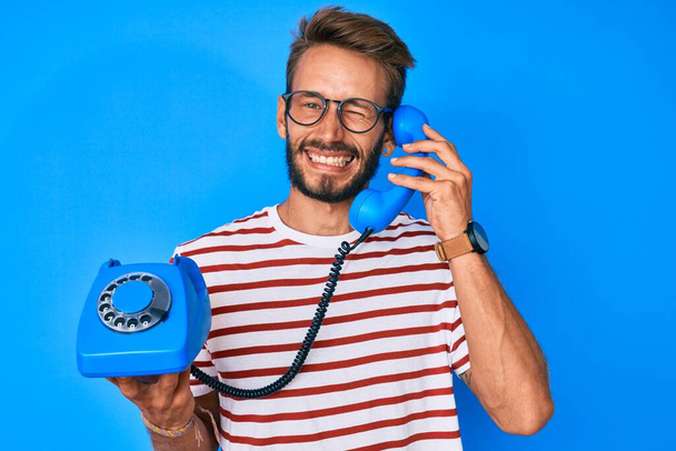 Όμορφος καυκάσιος άνδρας με γενειάδα κρατώντας vintage τηλέφωνο να κλείνει το μάτι στην κάμερα με σέξι έκφραση, χαρούμενο και χαρούμενο πρόσωπο.  - Φωτογραφία, εικόνα
