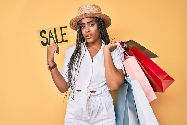 Młoda Afroamerykanka z warkoczami trzymająca torby na zakupy i baner sprzedażowy bez pojęcia i zdezorientowana ekspresja. koncepcja wątpliwości.  - Zdjęcie, obraz