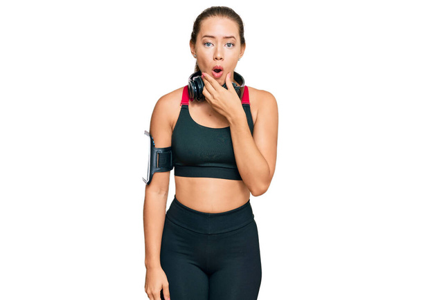 Beautiful blonde woman wearing gym clothes and using headphones looking fascinated with disbelief, surprise and amazed expression with hands on chin  - Photo, Image