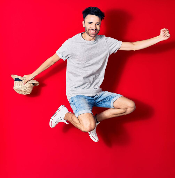 Jonge knappe Latijns-Amerikaanse man met casual kleding die vrolijk lacht. Springen met een glimlach op het gezicht met de zomer hoed vieren met vuist omhoog over geïsoleerde rode achtergrond. - Foto, afbeelding