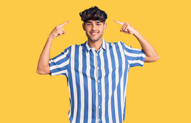 Gündelik kıyafetler giyen genç İspanyol adam gülümseyerek iki eliyle kafasını işaret ediyor. Harika bir fikir ya da düşünce, iyi bir hafıza.  - Fotoğraf, Görsel
