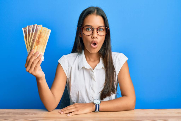 Beautiful hispanic woman holding 500 norwegian krone banknotes scared and amazed with open mouth for surprise, disbelief face  - Photo, Image