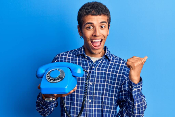 Fiatal afro-amerikai férfi kezében vintage telefon mutatva hüvelykujját fel az oldalon mosolyogva boldog nyitott szájjal  - Fotó, kép