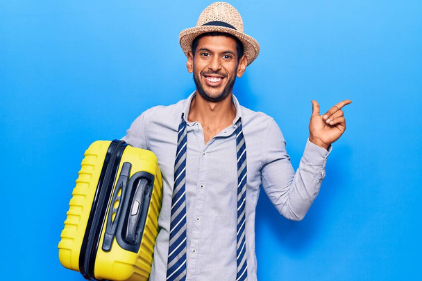 Joven hombre latino con sombrero de verano sosteniendo bolsa de cabina sonriendo feliz señalando con la mano y el dedo a un lado  - Foto, imagen