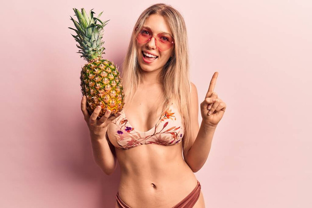 Mladá krásná blondýnka v bikinách a slunečních brýlích s ananasem s úsměvem s nápadem nebo otázkou ukazující prstem se šťastnou tváří, číslo jedna  - Fotografie, Obrázek