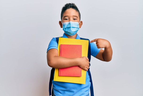 Kleiner hispanischer Junge mit medizinischer Maske geht zur Schule und lächelt glücklich und zeigt mit Hand und Finger  - Foto, Bild