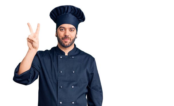 Mladý hispánec v kuchařské uniformě ukazující a ukazující prsty číslo dvě a usmívající se sebevědomě a šťastně.  - Fotografie, Obrázek