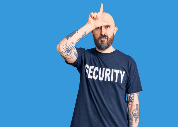 Молодой красивый мужчина в футболке с надписью "Охрана" высмеивает людей с пальцами на лбу, высмеивая и оскорбляя жесты неудачников..  - Фото, изображение
