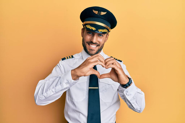 Przystojny Hiszpan w mundurze pilota samolotu, uśmiechnięty zakochany, robiący dłońmi symbole serca. romantyczna koncepcja.  - Zdjęcie, obraz