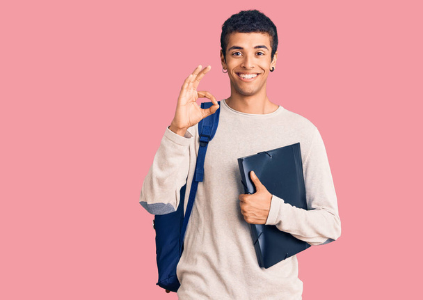 Jonge Afrikaanse amerikaanse man draagt student rugzak met bindmiddel doet ok teken met de vingers, glimlachend vriendelijk gebaar uitstekend symbool  - Foto, afbeelding