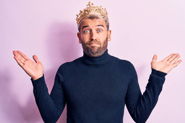 Young handsome blond man with piercing wearing golden king crown over pink background clueless and confused with open arms, no idea and doubtful face. - Fotoğraf, Görsel