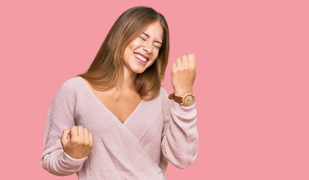 カジュアルな冬のピンクのセーターを着て美しいブロンドの女性は、腕を上げて目を閉じて成功に驚いて驚いています。勝者のコンセプト.  - 写真・画像