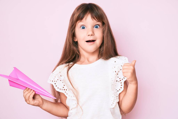 Little kaukaskie dziecko dziewczyna z długimi włosami trzymając papierowy samolot wskazując kciuk w górę do boku uśmiechając się szczęśliwy z otwartymi ustami  - Zdjęcie, obraz