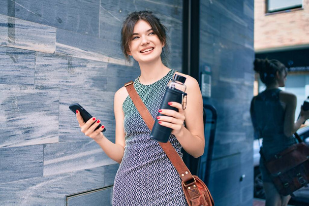 Giovane donna d'affari caucasica utilizzando smartphone in possesso di bottiglia d'acqua in città. - Foto, immagini