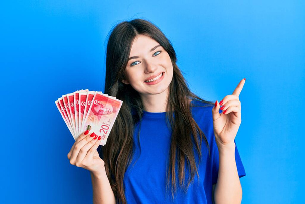 młoda piękne kaukaskie dziewczyna gospodarstwa 20 israel sykle banknoty uśmiechając szczęśliwy wskazując z ręki i palca w bok  - Zdjęcie, obraz