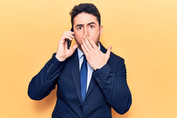 Joven hombre hispano vestido de traje conversando en el smartphone cubriendo la boca con la mano, sorprendido y temeroso de equivocarse. expresión sorprendida  - Foto, Imagen