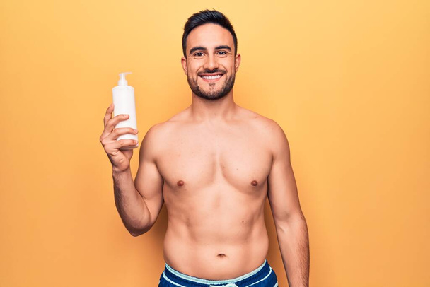 Schöner Mann mit Bart im Urlaub in Badebekleidung mit einer Flasche Sonnencreme im Stehen und glücklich lächelnd mit einem selbstbewussten Lächeln, das Zähne zeigt - Foto, Bild