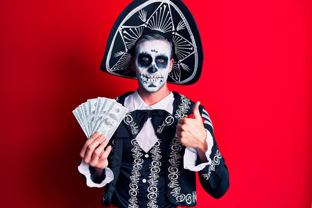 Young man wearing mexican day of the dead costume holding dollars smiling happy and positive, thumb up doing excellent and approval sign  - Φωτογραφία, εικόνα