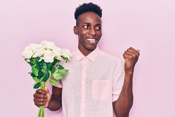 Mladý africký Američan drží květiny ukazující palcem nahoru na stranu s úsměvem šťastný s otevřenými ústy  - Fotografie, Obrázek
